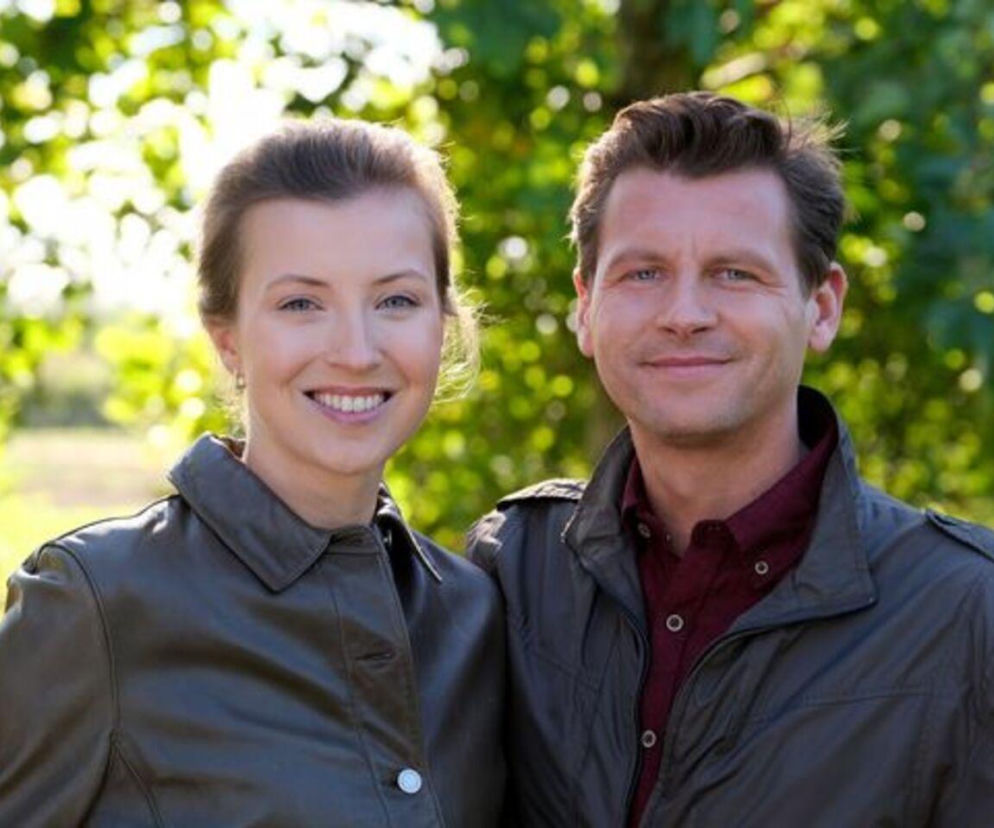 Dominika Suchecka jako Natalka i Patryk Szwichtenberg jako Adam Karski uśmiechają się do obiektywu, otoczeni zielenią. Para z serialu M jak miłość rozważa wspólne zamieszkanie. Więcej informacji o ich losach znajdziesz na stronie Super Seriale.