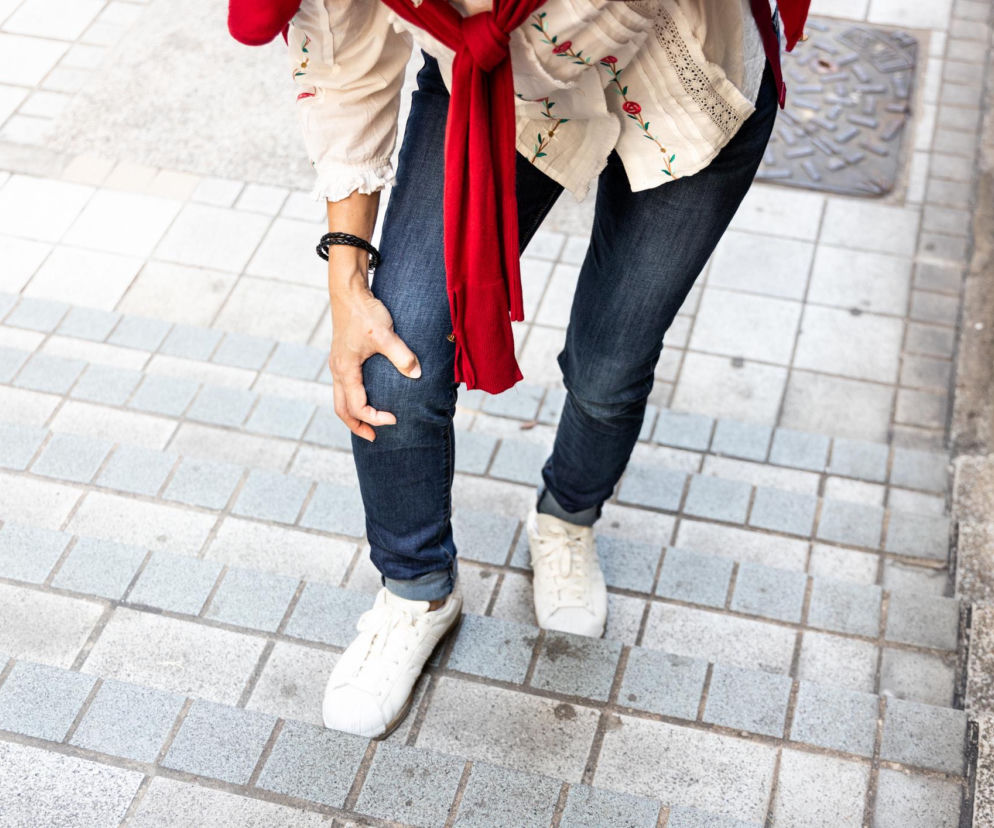 Kobieta w dżinsach i białych sneakersach, z czerwoną bluzą przewiązaną w pasie, stojąca na schodach i trzymająca się za bolące kolano. Artykuł na Poradnik Zdrowie wyjaśnia, jak chodzenie tyłem po schodach może pomóc złagodzić ból kolan.