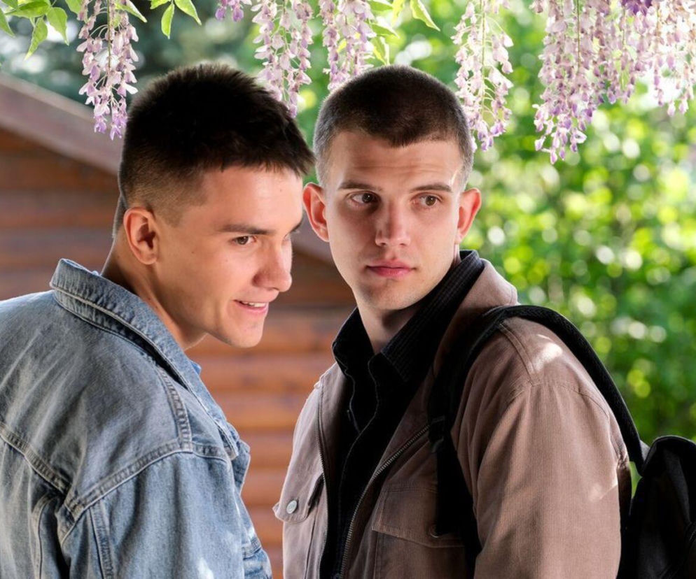 Dwóch młodych mężczyzn, Mateusz (Rafał Kowalski) w jeansowej kurtce i Janek (Jakub Sirko) z plecakiem, rozmawia na tle drewnianego domu i kwitnącej wisterii. Ich interakcja oddaje delikatną sprawę rodzinną z odcinka M jak miłość, o której możesz przeczytać na Super Seriale.