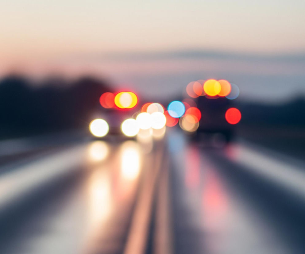 Rozmyte światła samochodów jadących w obu kierunkach dominują na obrazie, tworząc efekt bokeh na drodze, która jest odbija światło. Horyzont ukazuje odcienie różu, błękitu i szarości zachodzącego słońca, kontrastując z ciemnymi, niewyraźnymi sylwetkami drzew po bokach drogi. Jasne, białe reflektory pojazdów po lewej stronie drogi oraz czerwone i żółte tylne światła po prawej, rozlewają się w długie smugi światła na nawierzchni.