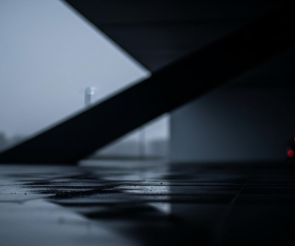 Na pierwszym planie, wilgotna, ciemna powierzchnia odbija światło, tworząc lśniące refleksy. Po lewej stronie widać jasno świecącą lampę uliczną na słupie, emitującą niebieskawą poświatę w szarej mgle. Po prawej stronie w oddali widoczne są trzy rozmyte, czerwone światła, prawdopodobnie lampy tylne pojazdu, których odbicia również rozciągają się na mokrej nawierzchni. Na drugim planie dominują ciemne, geometryczne kształty architektury, przypominające schody lub rampę, zanikające w zamglonym tle.