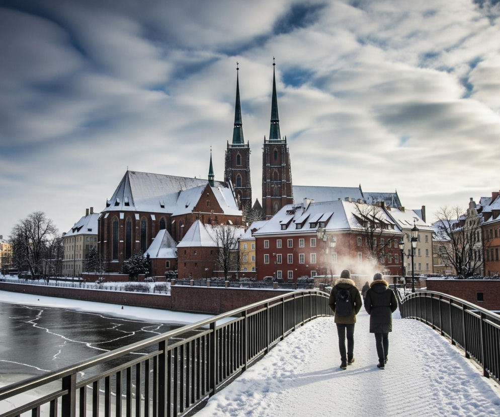 Dwie osoby idą po zasypanym śniegiem moście, oddalając się od obserwatora, z widokiem na zamarzniętą rzekę i panoramę miasta w tle. Most z metalową balustradą prowadzi nad rzeką, której brzegi pokryte są śniegiem i lodem, a powierzchnia wody jest częściowo zamarznięta z widocznymi pęknięciami lodu tworzącymi wzory. W tle dominują historyczne budynki z czerwonej cegły, w tym duży kościół z dwoma ostrymi wieżami, których dachy również pokryte są śniegiem. Niebo jest częściowo zachmurzone, z wyraźnym ruchem chmur widocznym w formie długich smug, co sugeruje długi czas naświetlania.