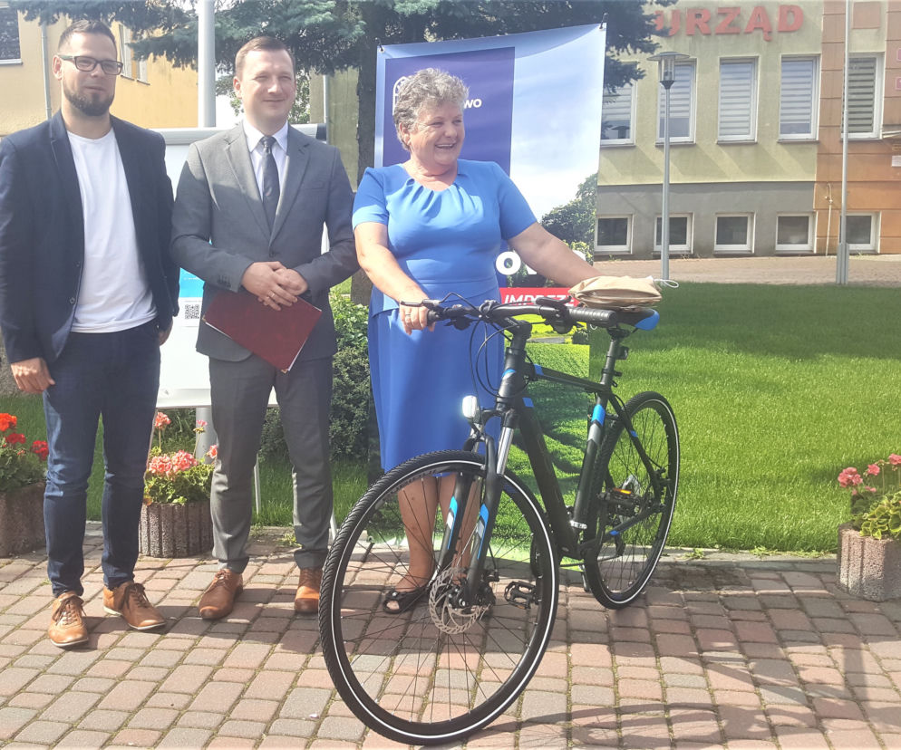 Dwaj mężczyźni i uśmiechnięta kobieta z rowerem przed budynkiem urzędu. O zmianach w meldunku przeczytasz na SE Superbiz.