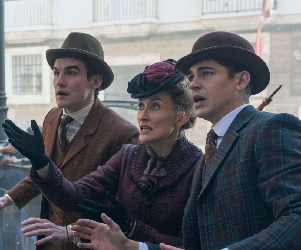 Hero Fiennes Tiffin i Max Irons w kapeluszach wraz z aktorką w bordowym kapeluszu i żakiecie, wszyscy z przerażeniem patrzący w jednym kierunku, z otwartymi ustami i uniesionymi dłońmi, w scenie z serialu Młody Sherlock. Więcej o nowej produkcji przeczytasz na Super Seriale.