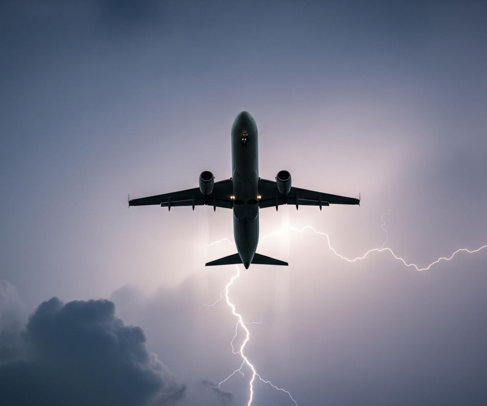 Samolot pasażerski, widziany od spodu, leci prosto w górę w kierunku centrum kadru, jego sylwetka jest ciemna na tle jasnego błysku pioruna. Skrzydła samolotu są rozłożone symetrycznie, a pod nimi widać dwa silniki i delikatne światła lądowania. Jasny, zygzakowaty piorun rozświetla fioletowo-szare niebo, rozchodząc się od środka ku prawemu dolnemu rogowi, z innym odgałęzieniem sięgającym ku górze po prawej. Po lewej stronie widoczne są ciemniejsze, puszyste chmury, które dodają dramatyzmu scenie burzy.