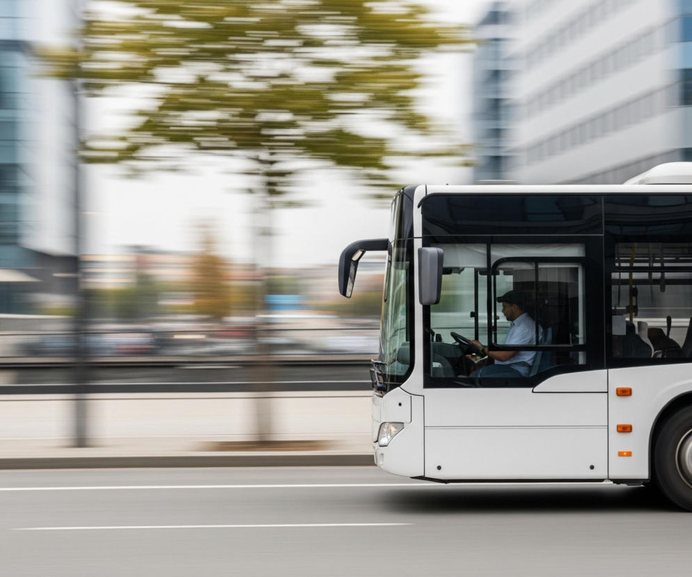 Biały autobus miejski porusza się szybko po szarej jezdni, na której widoczna jest jedna biała linia. Wewnątrz autobusu, za kierownicą, siedzi mężczyzna w jasnej koszuli i czapce. Tło rozmazane jest horyzontalnie, ukazując zielone liście drzew i szare, nowoczesne budynki z dużymi oknami.