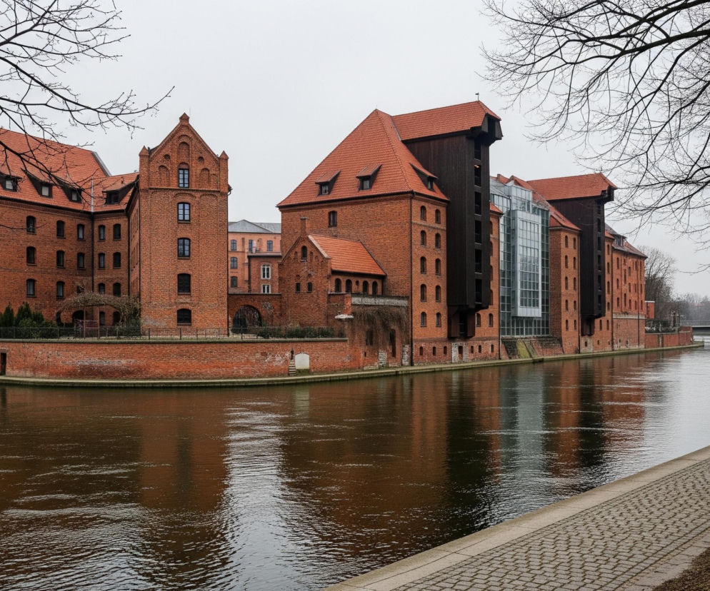 Na pierwszym planie, z prawej strony, widoczna jest postać osoby idącej tyłem, ubrana w jasną, puchową kurtkę z futrzanym kołnierzem i szarą czapkę, poruszająca się po brukowanej ścieżce biegnącej wzdłuż rzeki. Rzeka zajmuje większą część dolnej połowy kadru, jej ciemnobrązowa woda odbija światło i elewacje budynków po drugiej stronie. Po lewej stronie rzeki wznosi się kompleks zabytkowych budynków z czerwonej cegły, z licznymi oknami i spadzistymi dachami pokrytymi czerwoną dachówką. Budynki charakteryzują się zróżnicowaną architekturą, w tym szpiczastymi frontonami i elementami szachulcowymi, a także nowoczesną szklaną fasadą w jednym z nich. Nagie gałęzie drzew, pozbawione liści, oplatają kadr od góry, częściowo przesłaniając szare, zachmurzone niebo.