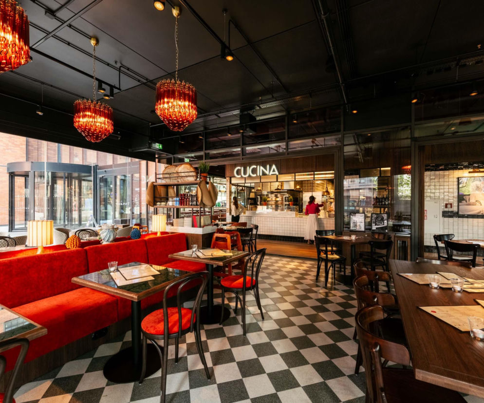 Wnętrze restauracji L'Osteria z czerwonymi kanapami i krzesłami, drewnianymi stołami oraz czarno-białą szachownicą na podłodze, podkreślające autentyczny włoski styl i atmosferę la dolce vita. Na końcu widać część kuchenną z napisem CUCINA i pracującym personelem. Więcej o ofercie na Dzień Ojca znajdziesz na naszym portalu.