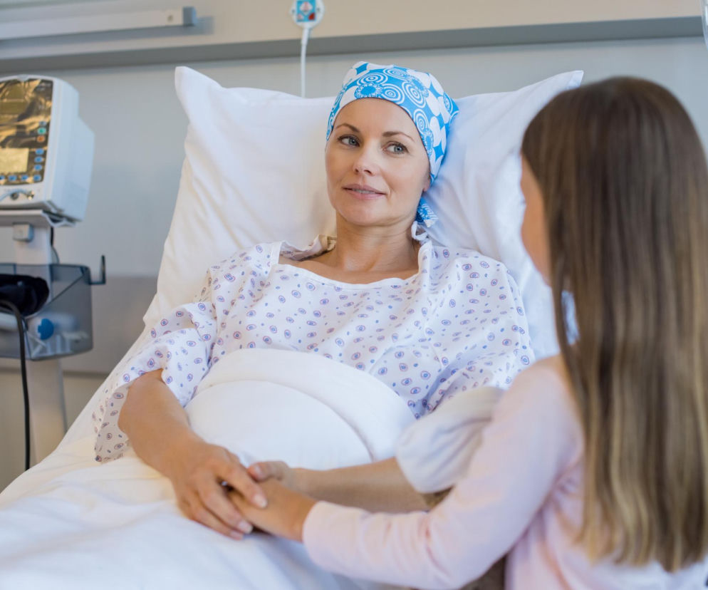 Pacjentka w szpitalnym łóżku, z niebiesko-białą chustą na głowie, trzyma za rękę dziewczynkę, symbolizując wsparcie w walce z chorobą nowotworową. O zwiększającej się liczbie zachorowań na raka, zwłaszcza w Wielkopolsce, przeczytasz na Poradnik Zdrowie.