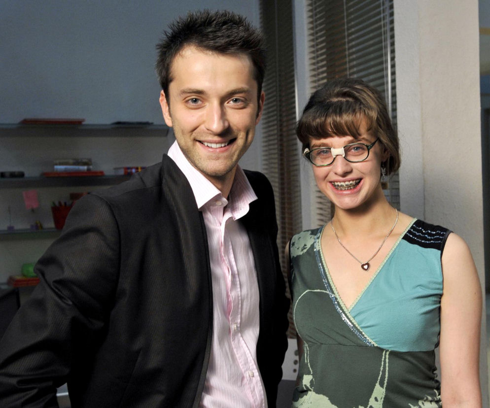 Ula Cieplak (Julia Kamińska) w okularach i aparacie ortodontycznym uśmiecha się obok Marka Dobrzańskiego (Filip Bobek) w garniturze. Zdjęcie z serialu Brzydula przedstawia głównych aktorów, o których metamorfozach przeczytasz na naszym portalu.