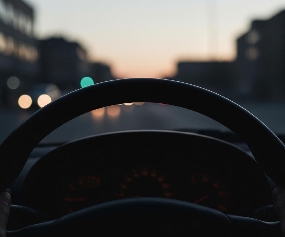 Dłonie trzymają czarną kierownicę samochodu, widoczną w dolnej części kadru. W tle, za deską rozdzielczą, rozciąga się miejski krajobraz o zmierzchu, z rozmazanymi światłami samochodów i budynków. Dominują ciemne odcienie szarości i błękitu, z pojedynczymi, rozmytymi punktami światła o barwie żółtej, czerwonej i turkusowej, a także jasnym, pomarańczowo-różowym pasmem zachodzącego słońca na horyzoncie.