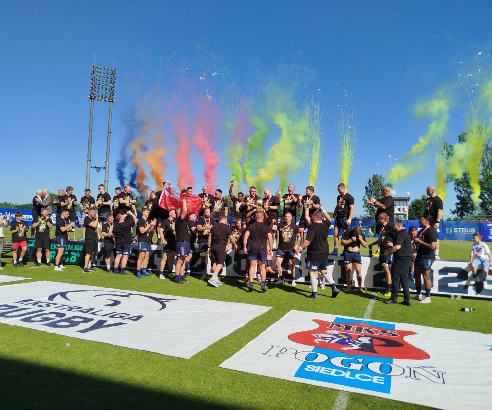 Drużyna MKS Awenta Pogoń Siedlce świętuje zwycięstwo na boisku rugby, trzymając trofeum i flagę Polski, otoczona kolorowym dymem i konfetti. To drugie z rzędu mistrzostwo Polski, o czym przeczytasz więcej na naszym portalu.
