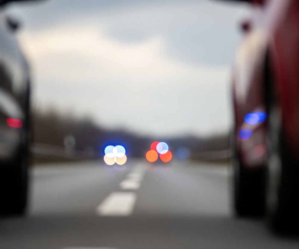 Dwa rozmyte samochody stoją na drodze, jeden po lewej stronie kadru, drugi po prawej, z widocznymi światłami bocznymi w kolorze czerwonym po lewej i niebieskim po prawej. Pomiędzy nimi rozciąga się szosa z widocznymi, białymi, przerywanymi liniami. W oddali, na środku jezdni, widać rozmazane światła: dwa niebieskie i dwa czerwone. Tło stanowi zamglone, jasne niebo, przechodzące w ciemniejszy, niewyraźny zarys drzew na horyzoncie.