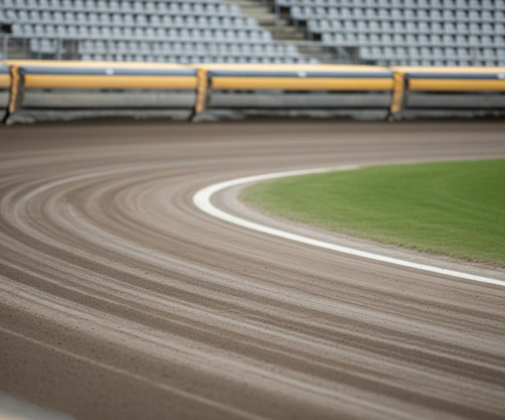 Tor żużlowy o brązowej, piaszczystej nawierzchni dominuje w kadrze, z wyraźnymi śladami przejeżdżających pojazdów, tworzącymi równoległe, ciemniejsze pasy. Krawędź wewnętrzna toru jest zaznaczona białą linią, za którą rozciąga się intensywnie zielony pas trawy. W tle widać rzędy pustych, szarych siedzeń stadionu, przed którymi biegnie żółta bariera ochronna.