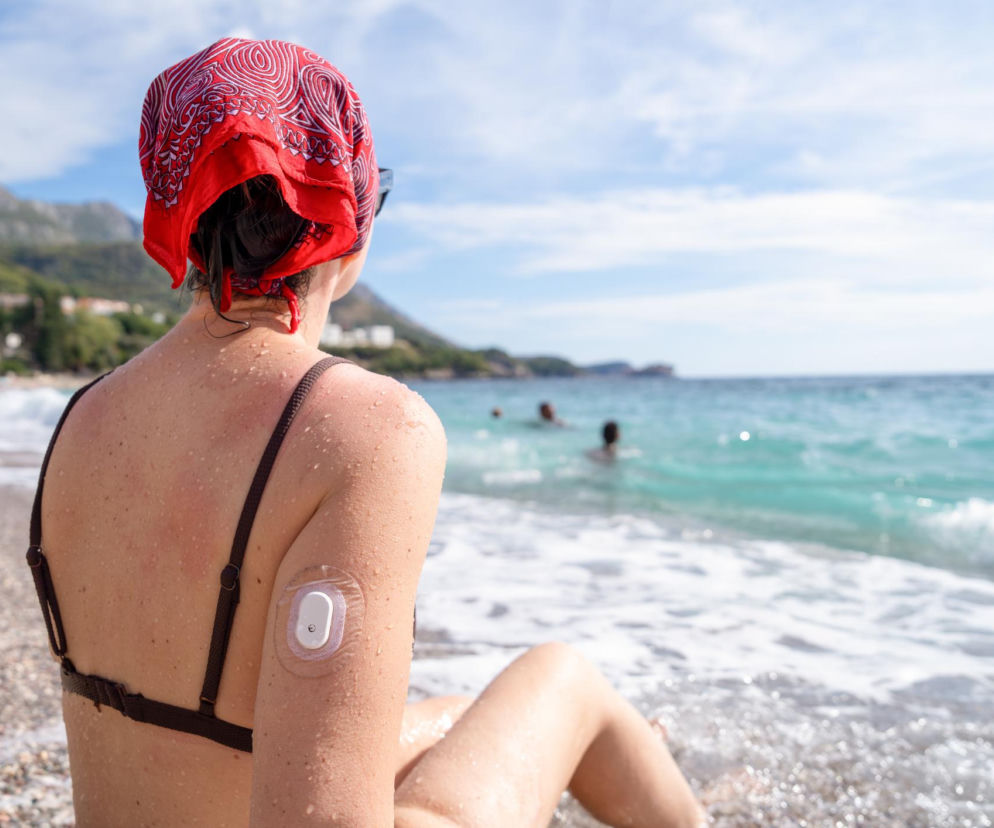 Kobieta w czerwonej chustce i kostiumie kąpielowym siedząca na plaży tyłem do obiektywu, obserwująca morze, z sensorem do ciągłego monitorowania glikemii na ramieniu. Zdjęcie ilustruje kontekst artykułu o cukrzycy typu 2 i jej monitorowaniu, o czym można przeczytać na Poradnik Zdrowie.