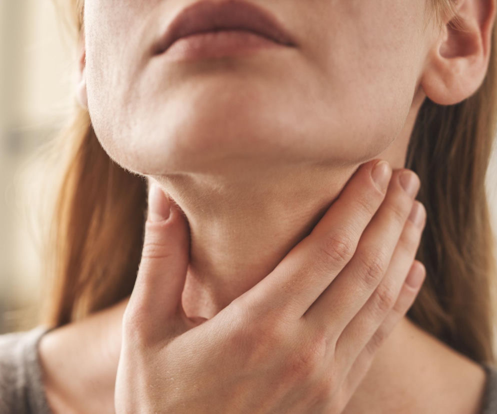 Blondynka w szarym swetrze trzyma dłoń na szyi, odczuwając dyskomfort związany z gulą w gardle. Problem może mieć związek z przeciążeniem kręgosłupa szyjnego, o czym szerzej przeczytasz na portalu Poradnik Zdrowie.