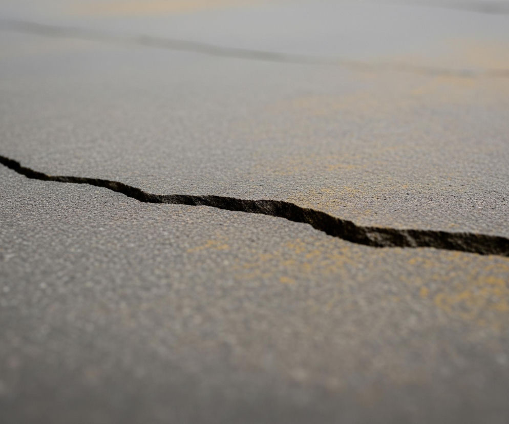 Przez środek szarej, asfaltowej powierzchni biegnie głębokie, ciemne pęknięcie, zaczynając z lewej strony kadru i kierując się w prawo, a następnie lekko w górę. Faktura powierzchni jest nierówna, ziarnista, z drobnymi kamykami i jaśniejszymi, żółtawymi przebarwieniami. Obiekt jest ostro zogniskowany na pierwszym planie, podczas gdy tło staje się coraz bardziej rozmyte.