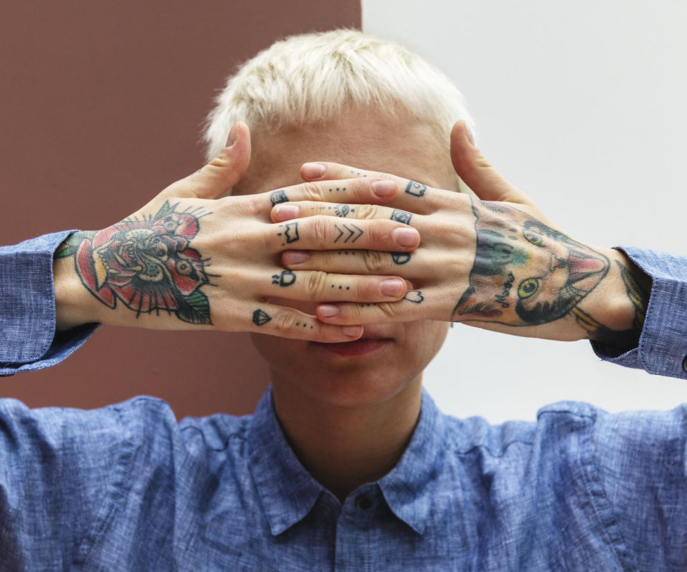 Osoba z krótkimi, blond włosami, w niebieskiej koszuli, zasłaniająca twarz wytatuowanymi dłońmi, symbolizuje obawę o wzrok po wykonaniu tatuażu, o czym można przeczytać na portalu Poradnik Zdrowie. Tatuowane dłonie z rysunkami kotów i róż podkreślają problem, że tusz może prowadzić do ślepoty.