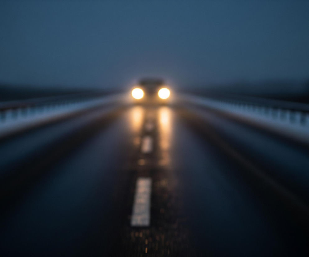 Rozmyta perspektywa ukazuje ciemną, asfaltową drogę biegnącą wprost w dal, oświetloną przez zbliżające się światła samochodu. Dwa jasne, żółtawe reflektory pojazdu są centralnym punktem kompozycji, rzucając wydłużone refleksy na mokrą nawierzchnię jezdni. Po obu stronach drogi widać niewyraźne, ciemne obiekty, prawdopodobnie barierki, a w tle unosi się mglista, granatowa mgła, która zaciera horyzont.
