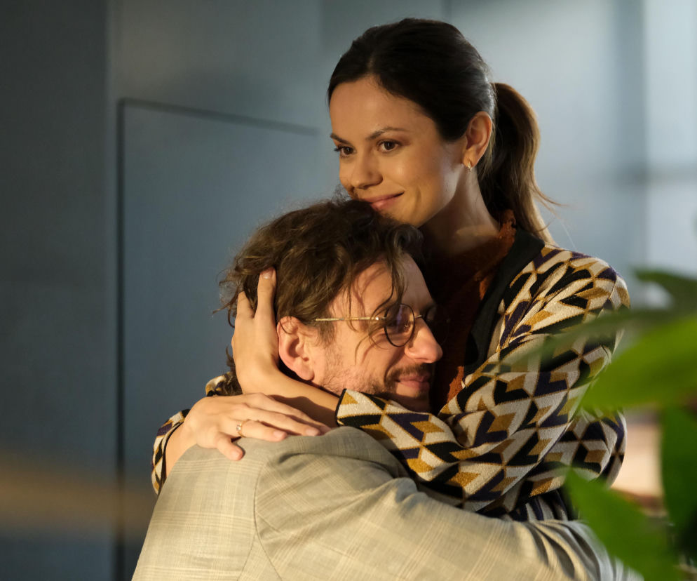 Paulina Lasota (Kasia) obejmująca Mateusza Mosiewicza (Mariusz) w scenie z serialu M jak miłość. Kasia z uśmiechem obejmuje Mariusza od tyłu, kładąc dłonie na jego ramionach, prezentując pierścionek zaręczynowy. Para, ubrana w casualowe stroje, ukazuje intymną chwilę. Więcej o ich losach na Super Seriale.