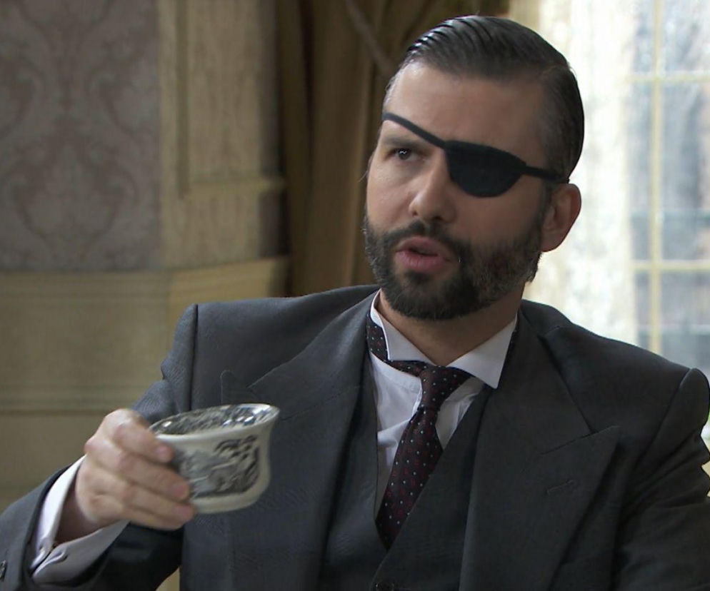 Mężczyzna z brodą i opaską na oku, ubrany w elegancki garnitur z kamizelką, trzyma ozdobną filiżankę. Postać, prawdopodobnie Jaime, mierzy się z problemami w związku z Ursulą i Blanką, o czym przeczytasz więcej na naszym portalu w streszczeniu odcinka 905 Akacjowej 38.