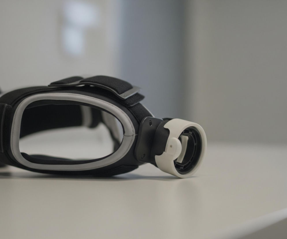 Na zdjęciu w centrum kadru leży czarno-białe urządzenie przypominające gogle, umieszczone na jasnej, płaskiej powierzchni. Urządzenie ma owalny kształt z dużym otworem pośrodku, obrzeżonym szarym materiałem, oraz czarną, teksturowaną powierzchnię zewnętrzną. Z prawej strony wystaje biały element, który ma w sobie okrągły, czarny detal z wystającymi elementami. Tło jest rozmyte, utrzymane w jasnych, neutralnych odcieniach szarości, z subtelnymi zmianami jasności, sugerującymi obecność okna lub źródła światła po prawej stronie.