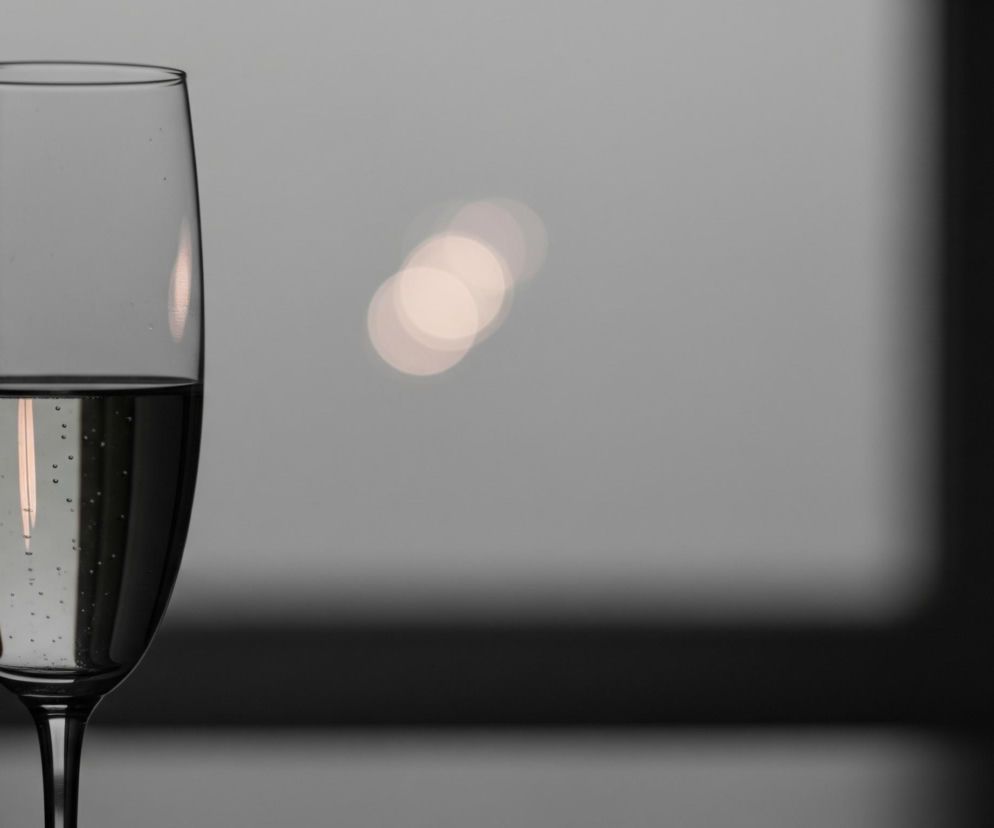 Wysoka szklanka na nóżce, wypełniona do połowy klarownym płynem, dominuje po lewej stronie kadru. W szklance widoczne są drobne bąbelki powietrza, a na jej powierzchni odbijają się jaśniejsze pionowe smugi światła. Tło jest rozmyte, zdominowane przez szarość, z trzema rozmytymi, jaśniejszymi okrągłymi plamami światła po prawej stronie, przypominającymi bokeh. Po prawej i na dole kadru widoczne są ciemniejsze, zamazane obszary, tworzące kontrast.