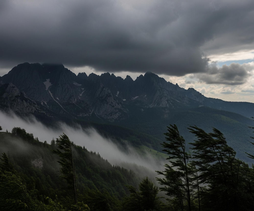 Górzysty krajobraz z dominującymi ciemnymi, poszarpanymi szczytami w oddali, częściowo zasłoniętymi przez gęste, ciężkie chmury, które pokrywają większą część nieba. Poniżej szczytów, gęste mgły spływają w dół zboczy, częściowo zakrywając zalesione pagórki w środkowej części obrazu. Na pierwszym planie widoczne są liściaste drzewa o ciemnozielonych koronach, których gałęzie i liście są wyraźnie pochylone w prawą stronę, co wskazuje na silny wiatr. Prawa górna część nieba ukazuje fragment błękitu z kilkoma jasnymi chmurami, kontrastującymi z ciemną, burzową atmosferą.