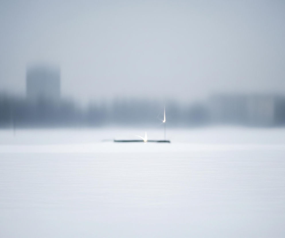Rozległy, pokryty śniegiem lub lodem teren, rozciągający się od pierwszego planu do horyzontu. W centrum kadru, na powierzchni, znajdują się dwa jasno świecące, pionowe płomienie lub iskry, przypominające zimne ognie, z których jeden jest lekko zakrzywiony. Płomienie wyrastają z ciemnego, podłużnego obiektu leżącego na śniegu. Tło jest zamglone i składa się z rozmazanych, szarych i granatowych sylwetek budynków lub drzew, sugerujących oddaloną linię zabudowy. Całość utrzymana jest w chłodnej, monochromatycznej palecie bieli, szarości i błękitu, co nadaje scenie zimowy, nieco tajemniczy charakter.