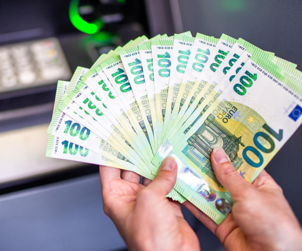 Osoba trzymająca w dłoniach rozłożone banknoty 100 euro, symbolizujące dużą sumę pieniędzy i inwestycje, z widocznym w tle bankomatem. Zdjęcie ilustruje temat miliardowych transakcji i rynkowych wyzwań, o których można przeczytać na Super Biznes.
