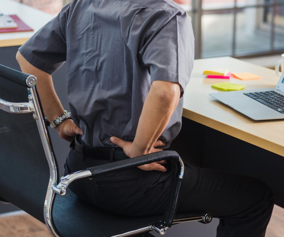 Mężczyzna w koszuli siedzący przy biurku z laptopem, trzymający się za obolałe plecy. Scena symbolizuje problem bólu kręgosłupa i syndromu martwego pośladka, o którym można przeczytać na portalu Poradnik Zdrowie.