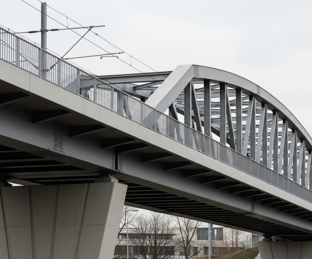 Masywny, nowoczesny most kolejowy wykonany z betonu i stali. Dominują odcienie szarości – od jasnego, niemal białego nieba, przez średnioszare elementy konstrukcji, po ciemniejsze cienie pod mostem. Górna część mostu, po której biegną tory, jest szara z widocznymi metalowymi barierkami oraz siecią trakcyjną i słupami. Jeden z żelbetowych filarów mostu o nieregularnym kształcie znajduje się po lewej stronie kadru, a drugi, nieco oddalony, po prawej, na tle nagich drzew i budynków o modernistycznej architekturze.