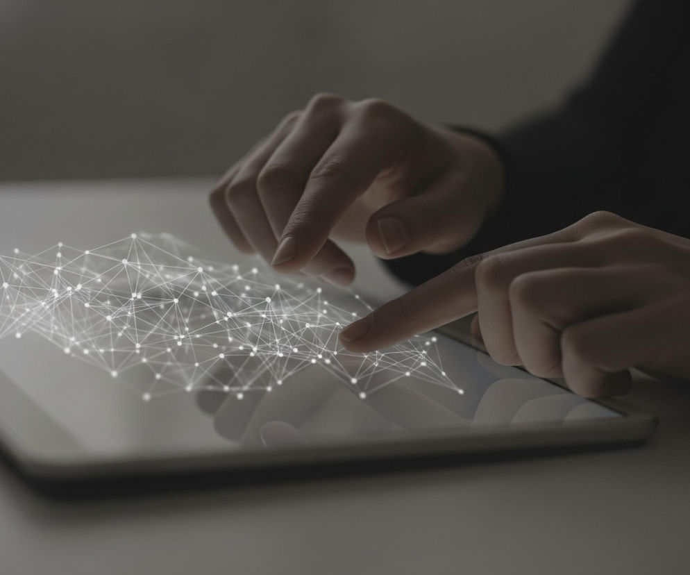 Dwie dłonie, należące do jednej osoby, obsługują tablet leżący na jasnym blacie. Na ekranie urządzenia, skierowanym ku górze, widoczna jest biała, cyfrowa grafika przedstawiająca połączone kropki i linie, tworzące siatkę. Palce lewej ręki lekko unoszą się nad ekranem, natomiast wskazujący palec prawej dłoni dotyka jego powierzchni. Tło jest ciemne, rozmyte, z niewyraźnie widocznym ciemnym materiałem odzieży w prawym górnym rogu.