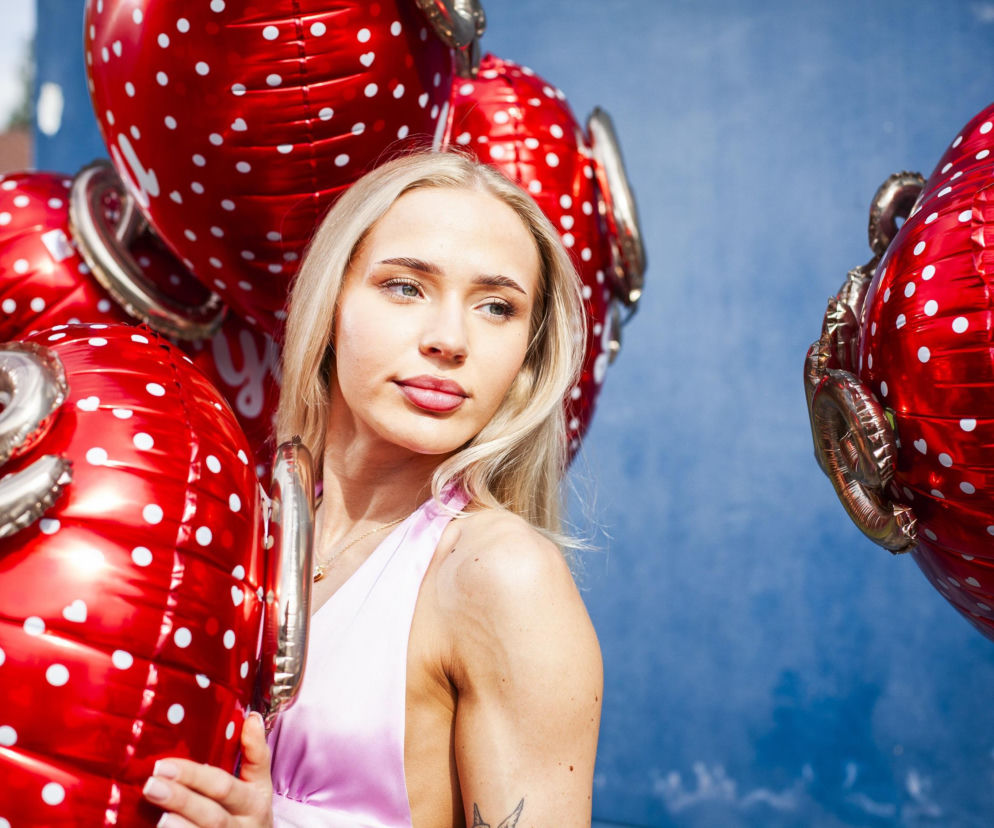 Uśmiechnięta Oliwka z blond włosami, ubrana w liliowy top, trzymająca czerwone balony w białe kropki, patrząca w prawo na tle niebieskiej ściany. Scena nawiązuje do intryg w programie Hotel Paradise, o których czytaj na Super Seriale.