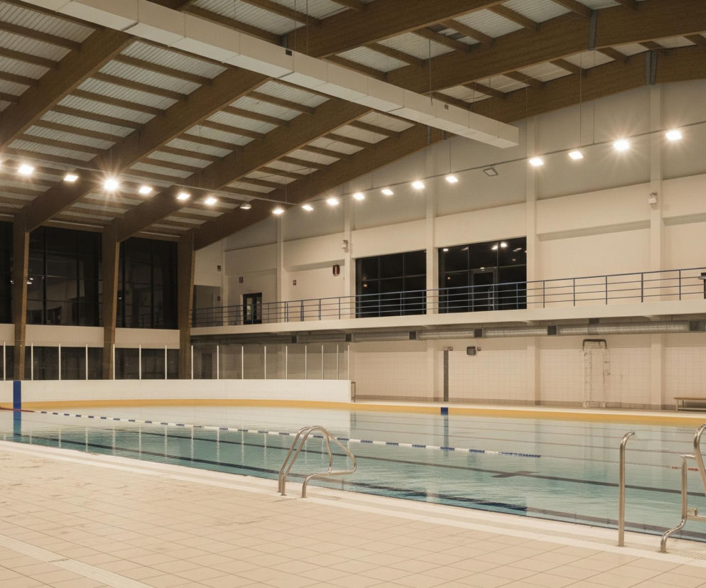 Wnętrze krytej pływalni z dużym basenem wypełnionym jasnoniebieską wodą zajmuje centralną część obrazu. Basen jest otoczony płytkami w beżowym odcieniu, a woda odbija światło z licznych lamp sufitowych. Nad basenem rozciąga się drewniany strop z białymi panelami, oświetlony rzędami kwadratowych lamp. Po prawej stronie basenu widoczne są metalowe barierki i ławki na podwyższeniu, natomiast lewa strona ukazuje ciemne okna i metalową ławkę na pierwszym planie.
