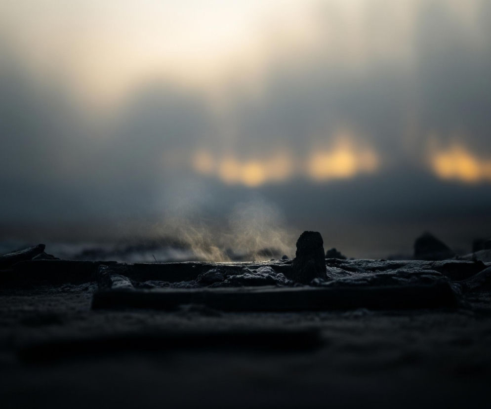 Na pierwszym planie widoczne są ciemne, nieregularne kształty, przypominające gruz lub zmarzniętą ziemię, z drobnymi, jasnymi akcentami sugerującymi pokrycie szronem lub pyłem. Z centralnej części tych kształtów unosi się delikatna smuga białej pary lub dymu, podświetlona z tyłu. Tło jest całkowicie rozmyte, zdominowane przez szaro-niebieską mgłę, przechodzącą w górnej części w jasny, żółto-pomarańczowy blask, prawdopodobnie słońca wschodzącego lub zachodzącego. Całość tworzy nastrojową scenę o chłodnych barwach, z kontrastującym ciepłym światłem w tle.