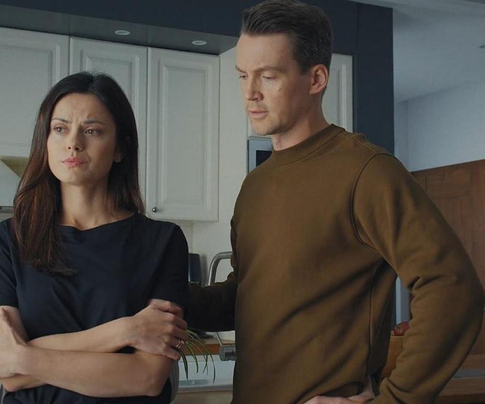 Paulina Lasota jako Kasia i Krzysztof Kwiatkowski jako Jakub w scenie z serialu M jak miłość. Kasia, z ramionami skrzyżowanymi, patrzy zamyślona, podczas gdy Jakub stoi obok z ręką na biodrze. Oboje znajdują się w kuchni, a ich spojrzenia oddają emocje związane z nadchodzącą prawdą o dziecku, o której można przeczytać na Super Seriale.