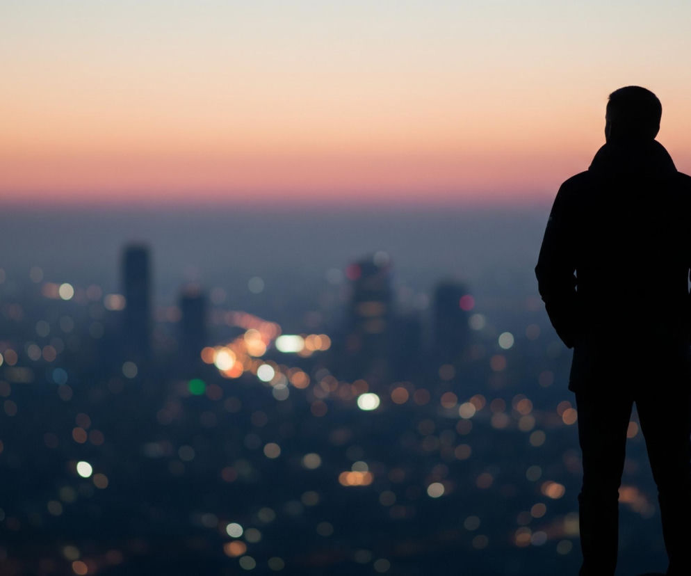 Mężczyzna w ciemnym ubraniu stoi tyłem do obserwatora, patrząc na rozległe, rozmazane światła miasta rozciągające się w oddali. Jego sylwetka jest widoczna jako czarny cień, umieszczony po prawej stronie kadru, na tle zachodzącego słońca, które stopniowo przechodzi od jasnego beżu i pomarańczy na górze do głębokiego różu i fioletu, a następnie granatu w kierunku horyzontu. Poniżej, w ciemnej mgle, majaczą niewyraźne kształty budynków i setki okrągłych, rozmytych punktów świetlnych, głównie w kolorach białym, żółtym i pomarańczowym, tworzących efekt bokeh.