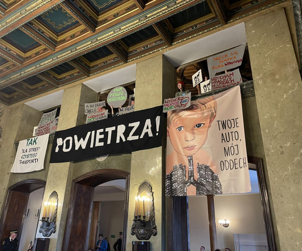 Wnętrze ozdobionej sali Radzie Miasta Krakowa, z której ludzie patrzą z balkonów, trzymając transparenty przeciwko Strefie Czystego Transportu. Widoczne hasła POWIETRZA!, TWOJE AUTO, MÓJ ODDECH oraz MAM PRAWO ODDYCHAĆ. Na dole sali widać pojedyncze osoby. Więcej o tej kwestii przeczytasz na Architektura Murator Plus.