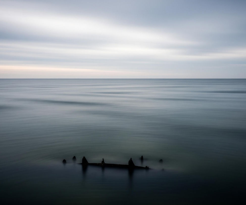 Ciemna, rozmyta konstrukcja o nieregularnych kształtach, z kilkoma wystającymi pionowymi elementami, spoczywa w dolnej części obrazu, częściowo zanurzona w spokojnej, ciemnogranatowej wodzie. Powierzchnia wody, lekko rozmyta, tworzy poziome, falujące smugi, rozciągające się aż po horyzont. Nad linią horyzontu rozciąga się zachmurzone niebo w odcieniach szarości i błękitu, z jaśniejszą, lekko różowawą smugą nisko nad horyzontem, tworząc rozmyty i spokojny pejzaż.