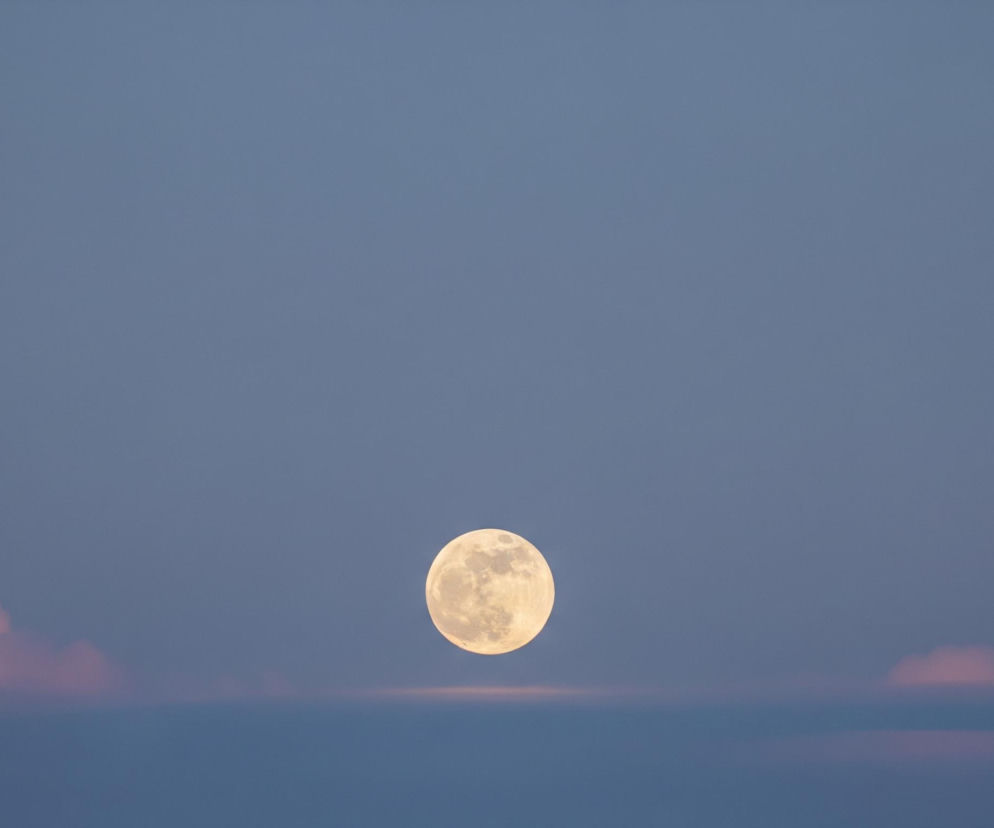 Pełnia księżyca dominuje centralną część obrazu, świecąc jasnym, żółtawo-białym blaskiem. Powierzchnia księżyca ma widoczne, ciemniejsze plamy, tworzące nieregularne wzory. Księżyc umieszczony jest na tle nieba o przejściowych odcieniach, z dominującym jasnoniebieskim kolorem u góry, przechodzącym w ciemniejszy, szaroniebieski odcień w dolnej części. Tuż pod księżycem, na horyzoncie, rozciąga się cienka, pozioma linia chmur, częściowo podświetlona od dołu na różowo-pomarańczowo. Dodatkowo, po lewej i prawej stronie tej linii, widoczne są pojedyncze, puszyste chmury, również zabarwione delikatnymi różowymi i pomarańczowymi tonami.