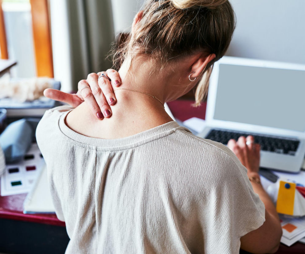 Kobieta masuje obolały kark, siedząc przy laptopie na biurku, co symbolizuje problem bólu karku od komputera, z którym zmaga się wielu pracowników biurowych. Dowiedz się, jak z nim walczyć na Poradnik Zdrowie.