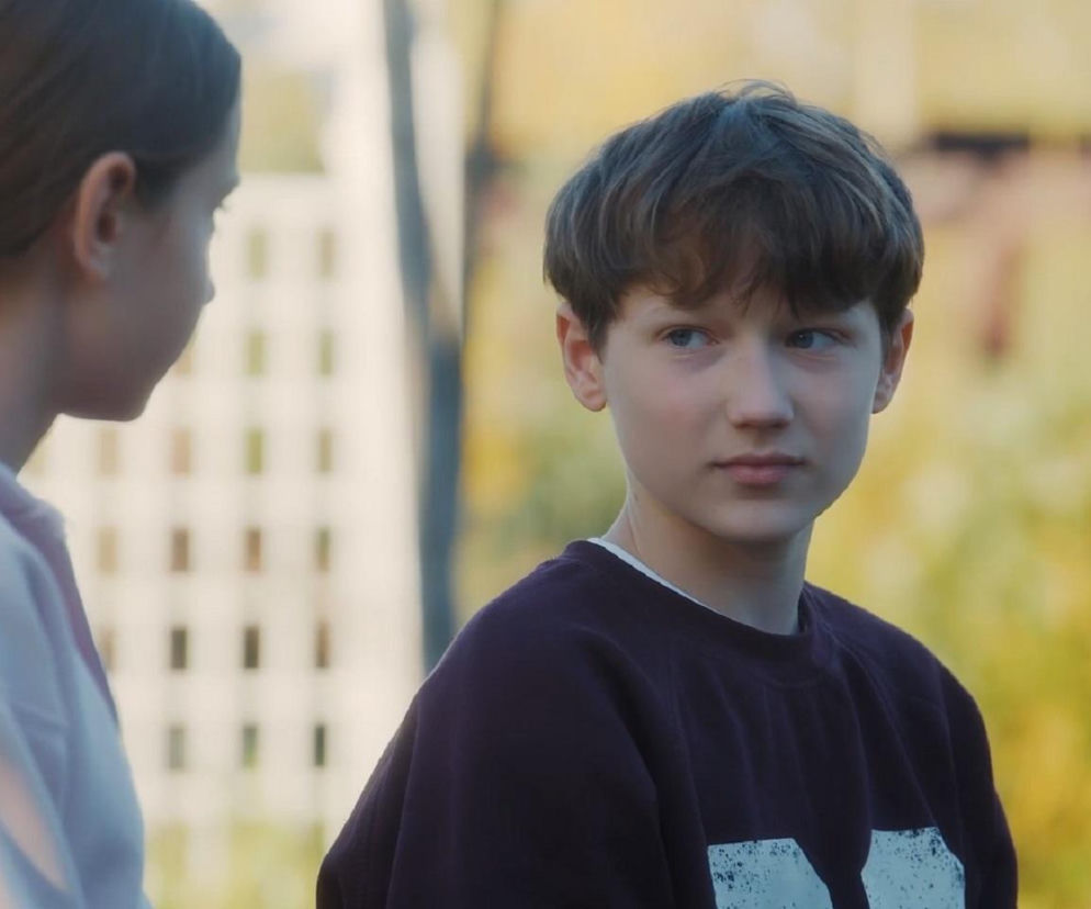 Ujęcie filmowe: młody chłopak Franek (Jarosław Śmigielski) w granatowej bluzie patrzy w lewo, z profilu widać Hanię (Wiktoria Basik) z warkoczem i w jasnej bluzie z kapturem; scena z serialu M jak miłość. Więcej o losach bohaterów przeczytasz na Super Seriale.