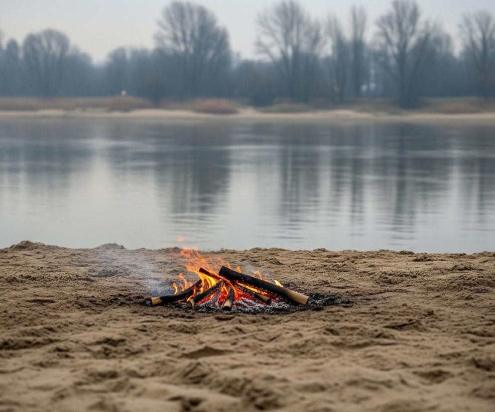 Na pierwszym planie, centralnie, pali się ognisko z kilku kawałków drewna ułożonych w stos na brązowym, piaszczystym podłożu, z widocznymi pomarańczowo-czerwonymi płomieniami i subtelnym dymem unoszącym się w lewą stronę. Za ogniskiem rozciąga się szeroki, spokojny zbiornik wodny w odcieniach szarości i błękitu, z lekkim zafalowaniem powierzchni. W tle, po drugiej stronie wody, widoczny jest odległy, rozmazany las z bezlistnymi drzewami o ciemnych, nieregularnych kształtach, zlewającymi się z bladym, szarym niebem.