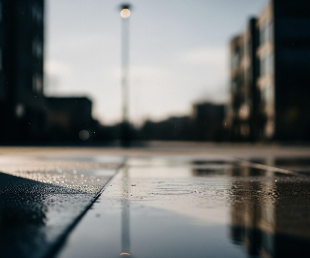 Powiększony fragment chodnika pokrytego drobnymi kroplami wody dominuje pierwszy plan, ukazując wyraźną fakturę powierzchni i odbijające się światło. Na dalszym planie, w centrum, widoczna jest rozmyta, jasna lampa uliczna na wysokim słupie, której światło odbija się w kałuży na dole kadru. Po obu stronach, w oddali, znajdują się ciemne, rozmazane sylwetki budynków z niewyraźnymi, jaśniejszymi elementami przypominającymi okna, a cała scena ma chłodny, stonowany odcień.