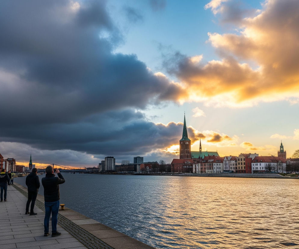 Scena przedstawia szeroki widok na rzekę, nabrzeże i panoramę miasta o zachodzie słońca. Duża połać ciemnoniebieskich chmur pokrywa lewą część nieba, przechodząc w jaśniejsze, złoto-pomarańczowe chmury po prawej stronie, oświetlone przez nisko położone słońce. Wzdłuż lewego brzegu rzeki rozciąga się promenada, na której idzie lub stoi kilka osób, niektóre z nich fotografują krajobraz telefonami komórkowymi. Po przeciwnej stronie rzeki widoczna jest zabudowa miejska z charakterystycznym wysokim, zielonym spiczastym dachem kościoła, otoczonym rzędem kolorowych kamienic i nowocześniejszych budynków. Złote światło odbija się od falującej tafli wody, tworząc świetliste smugi na jej powierzchni.