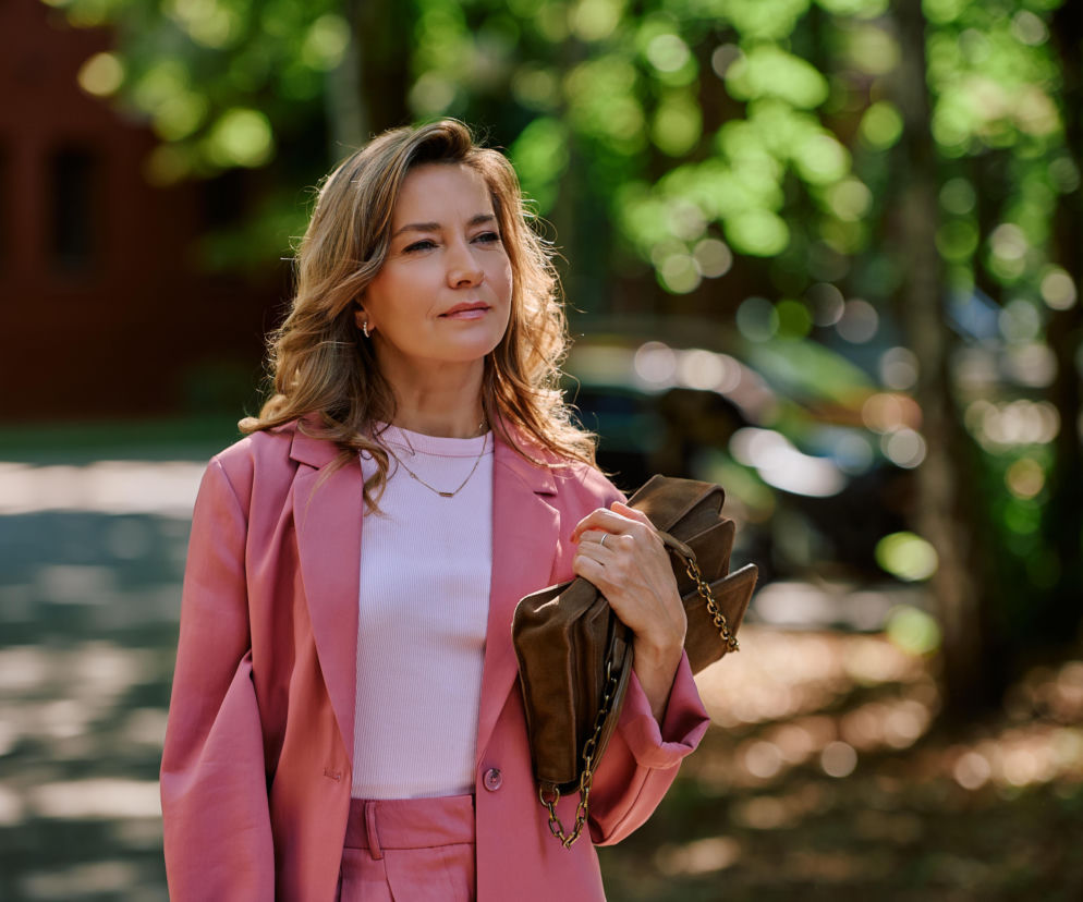 Kamila Baar jako Oriana Warska w różowym garniturze trzyma brązową torebkę. O jej roli w Na Wspólnej przeczytasz w Eska.