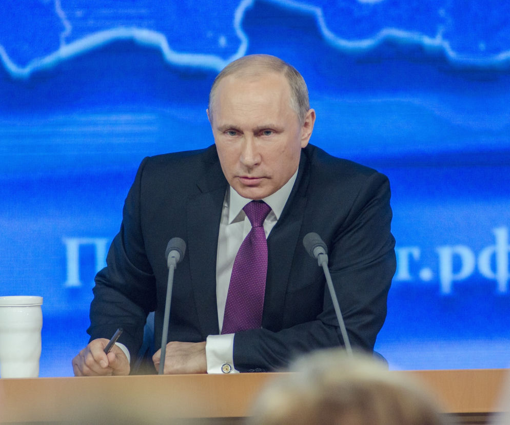 Prezydent Rosji Władimir Putin siedzi przy stole na tle niebieskiej mapy. O działaniach Rosji przeczytasz na SE.