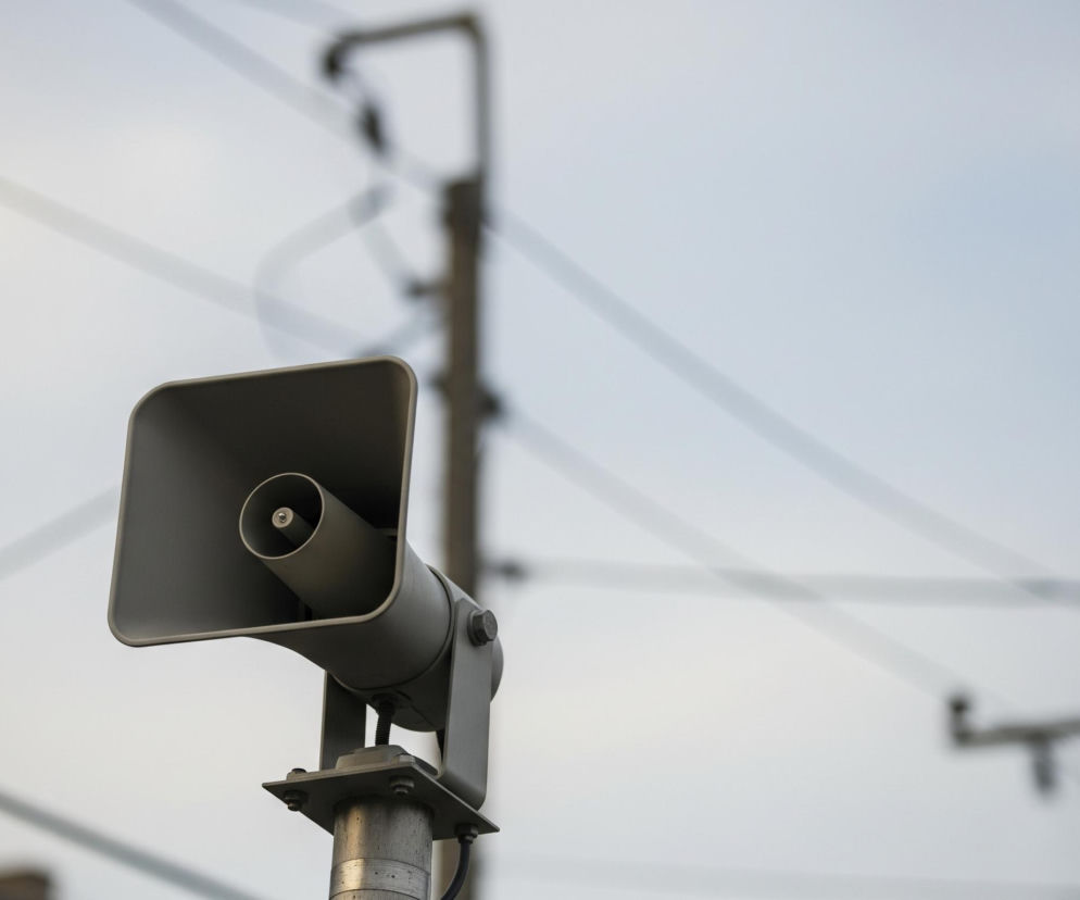 W centrum obrazu znajduje się szary megafon o prostokątnym kształcie z okrągłym wylotem, zamocowany na metalowym słupie. Megafon jest ustawiony pod lekkim kątem, skierowany w dół w prawą stronę. Tło stanowią rozmyte linie przypominające przewody elektryczne oraz niewyraźne sylwetki słupów energetycznych, wszystko na jasnym, zachmurzonym niebie.