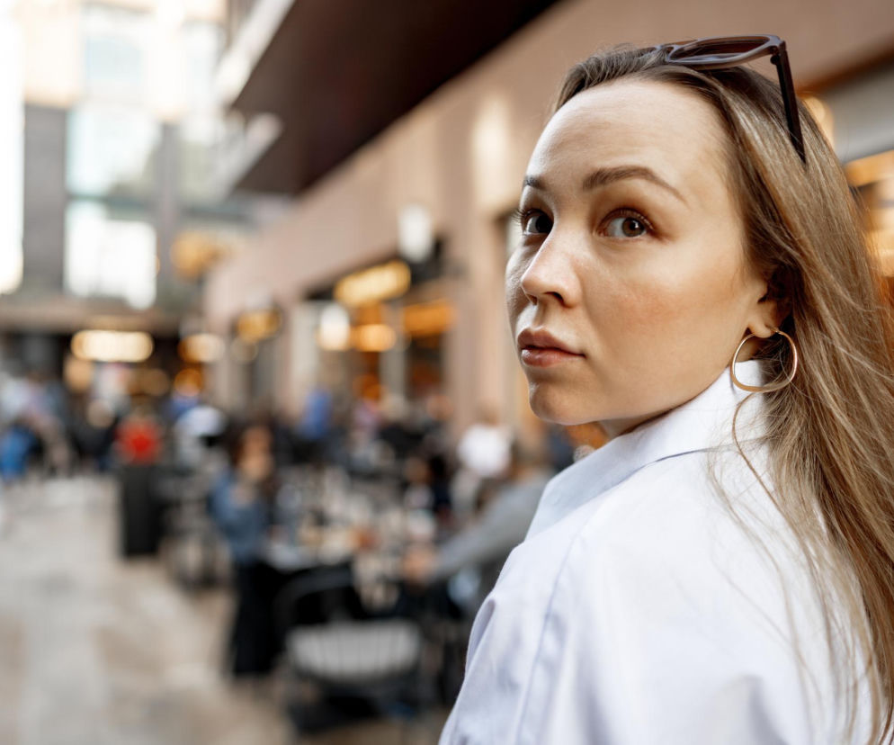 Kobieta zaniepokojona odwraca się przez ramię, patrząc na otoczenie, z okularami przeciwsłonecznymi na głowie. Jej wyraz twarzy sugeruje poczucie bycia obserwowaną, co może świadczyć o zaburzeniach lękowych, o czym możesz przeczytać na portalu Poradnik Zdrowie.
